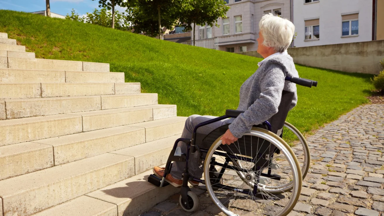 A person in a wheelchair looks up at a set of stairs outdoors, with grass and buildings in the background.