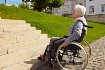 A person in a wheelchair looks up at a set of stairs outdoors, with grass and buildings in the background.