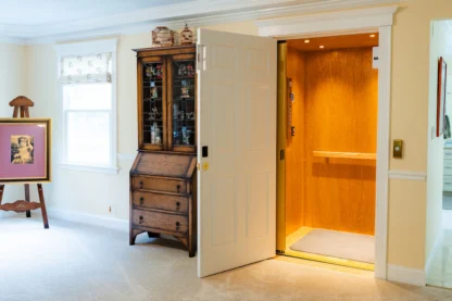 A residential interior with an open wooden elevator. A cabinet and framed artwork are nearby. Carpeted floor and white walls are visible.