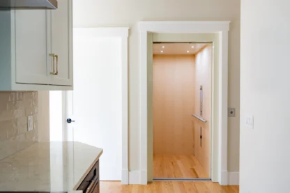 An open residential elevator with light wood interior, located next to a kitchen area with white cabinetry and light-colored walls.