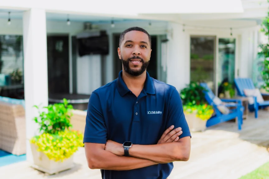 A man with a beard and short hair stands outdoors with arms crossed, wearing a blue polo shirt. There's patio furniture and greenery in the background.