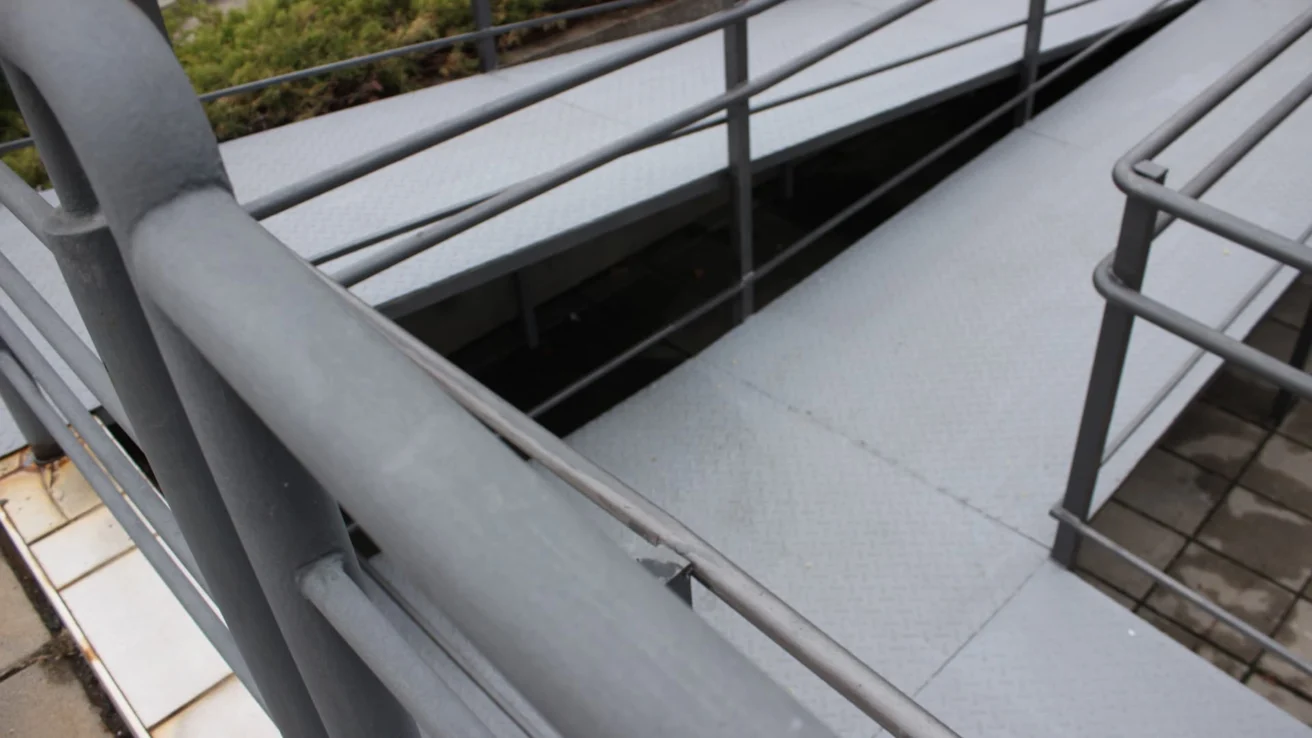 Close-up of a gray metal ramp with railings, leading downward. The surface is ridged, and shrubs are visible in the background.