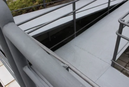 Close-up of a gray metal ramp with railings, leading downward. The surface is ridged, and shrubs are visible in the background.