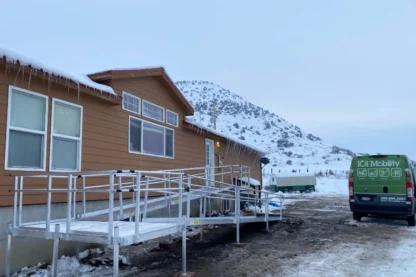 A snow-covered house with a metal accessibility ramp is in front of a mountain backdrop. A green van is parked nearby.