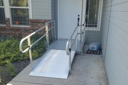 A metal wheelchair ramp leading to a doorway, with a welcome mat and a pack of water bottles on the porch.