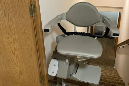 A gray stair lift chair installed on a staircase with beige carpeting and wooden side paneling.