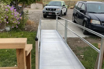 A metal wheelchair ramp leads from a wooden porch to a driveway with two parked vehicles. A tree and flowers are to the left.