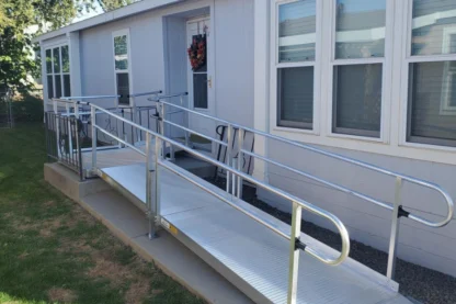 A metal wheelchair ramp leads to the entrance of a gray house, with windows and greenery visible.