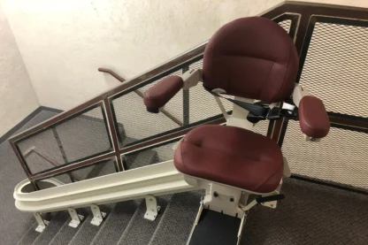 A stairlift with a burgundy seat is installed along a carpeted staircase with a metal railing.