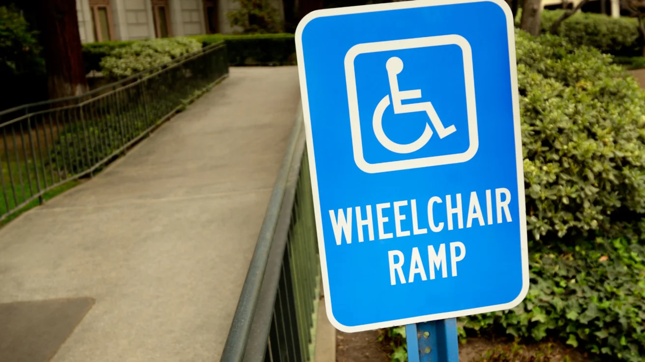 Blue sign with wheelchair symbol reads "Wheelchair Ramp" next to a sloped ramp leading to a building entrance surrounded by greenery.