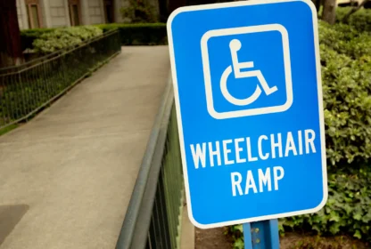 Blue sign with wheelchair symbol reads "Wheelchair Ramp" next to a sloped ramp leading to a building entrance surrounded by greenery.