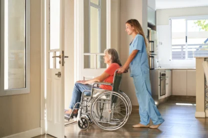 A caregiver in scrubs pushes an older woman in a wheelchair towards a sunlit doorway in a home setting.