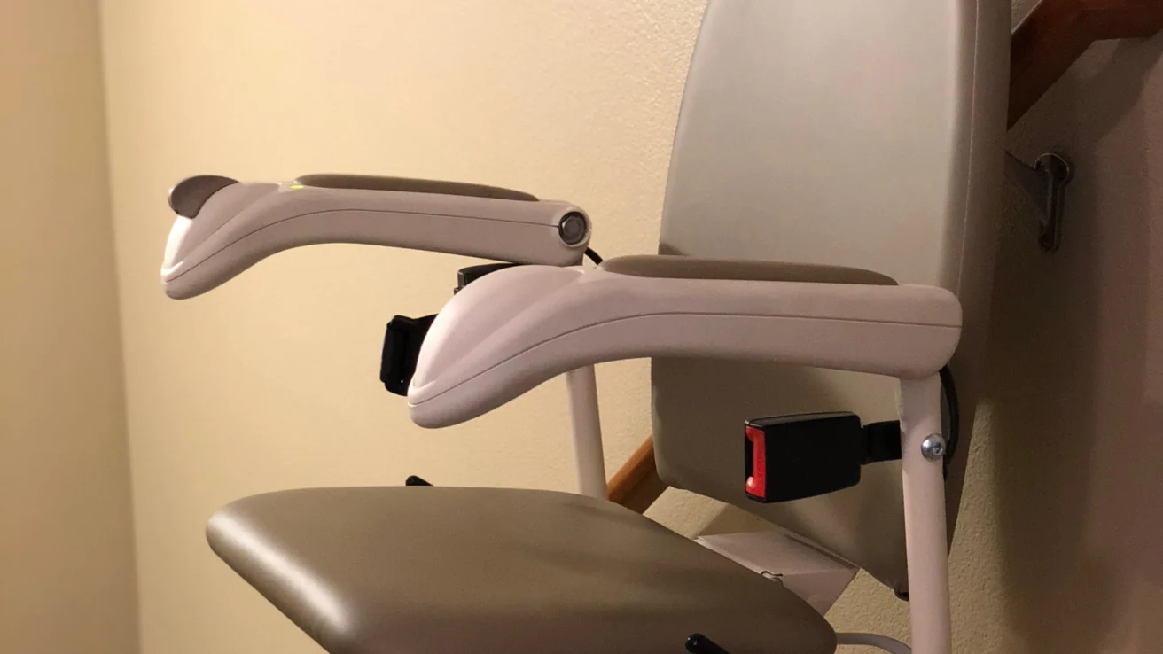 A beige stairlift chair with armrests and a safety belt is mounted on a staircase railing against a neutral wall.