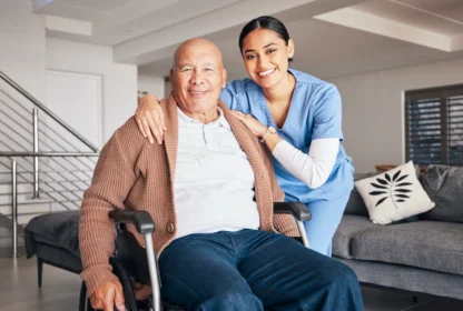 A smiling nurse stands beside an elderly man in a wheelchair in a living room.