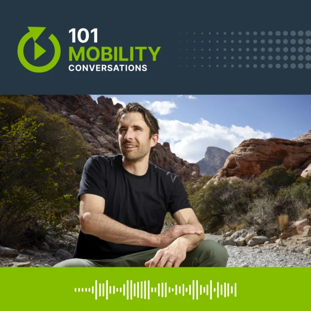 Man sitting outdoors in a rocky landscape with mountains and shrubs in the background, wearing a black t-shirt. Above, "101 Mobility Conversations" is written with a graphic and a waveform.