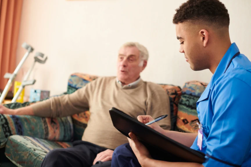 A healthcare worker in blue scrubs sits with an elderly man on a patterned sofa, taking notes on a clipboard. A pair of crutches rests against the wall in the background.