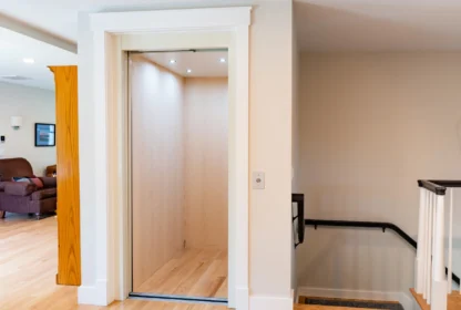 A residential elevator with an open door, light wood interior, and a control panel, situated beside a staircase with a black handrail.