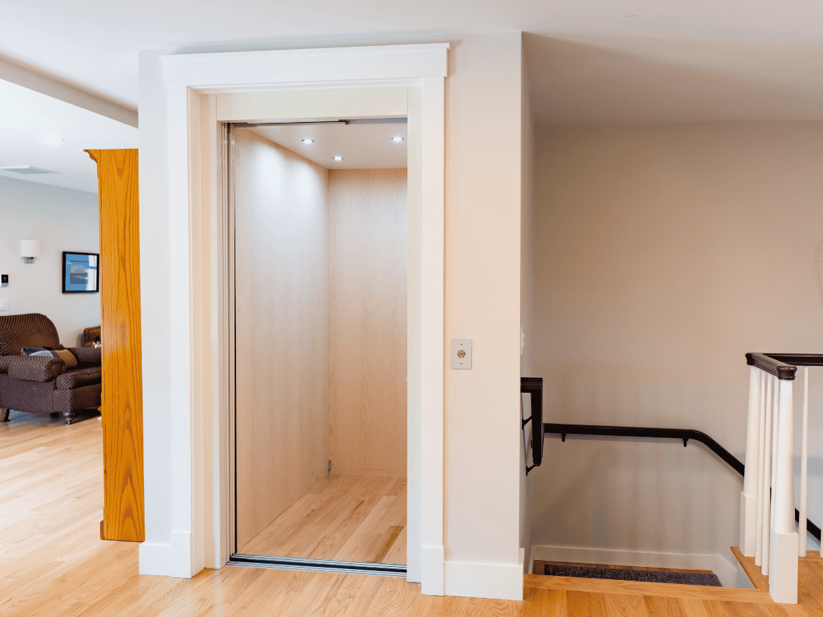 A residential elevator with an open door, light wood interior, and a control panel, situated beside a staircase with a black handrail.