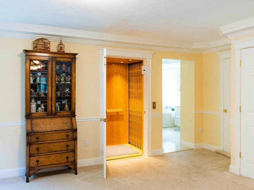 A wooden cabinet stands next to an open home elevator door in a warmly lit room with beige walls and carpet flooring.