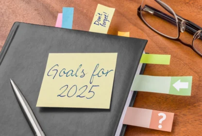 A planner with a "Goals for 2025" note on the cover, surrounded by colored tabs and a pair of glasses on a wooden desk.