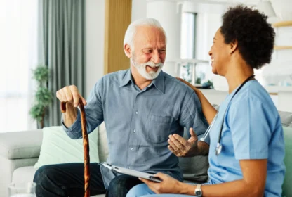 A smiling older man with a cane sits on a couch, talking to a healthcare professional in scrubs.